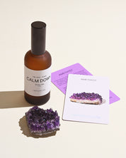 A bottle of 'Calm Down Room Mist' with a dark label and a cluster of amethyst crystals on a light background, alongside a purple card with text.