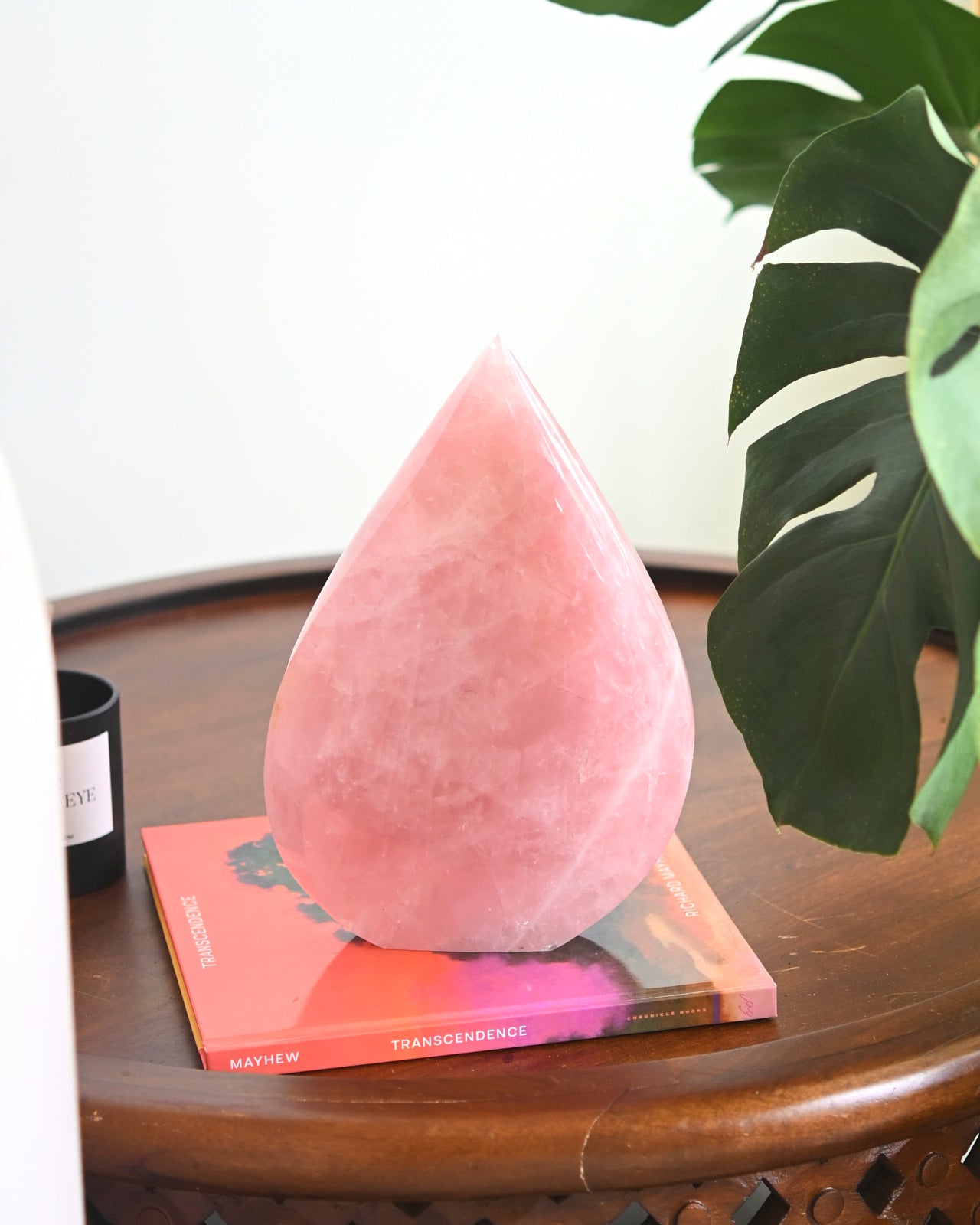 A large, pink Rose Quartz freeform crystal on top of a book on a wooden surface with a green plant in the background.