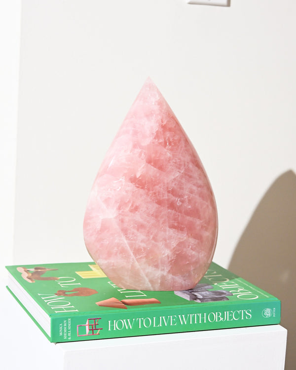 A large, pink, rose quartz freeform crystal weighing 5.1kg, placed on top of a book titled 'HOW TO LIVE WITH OBJECTS'.