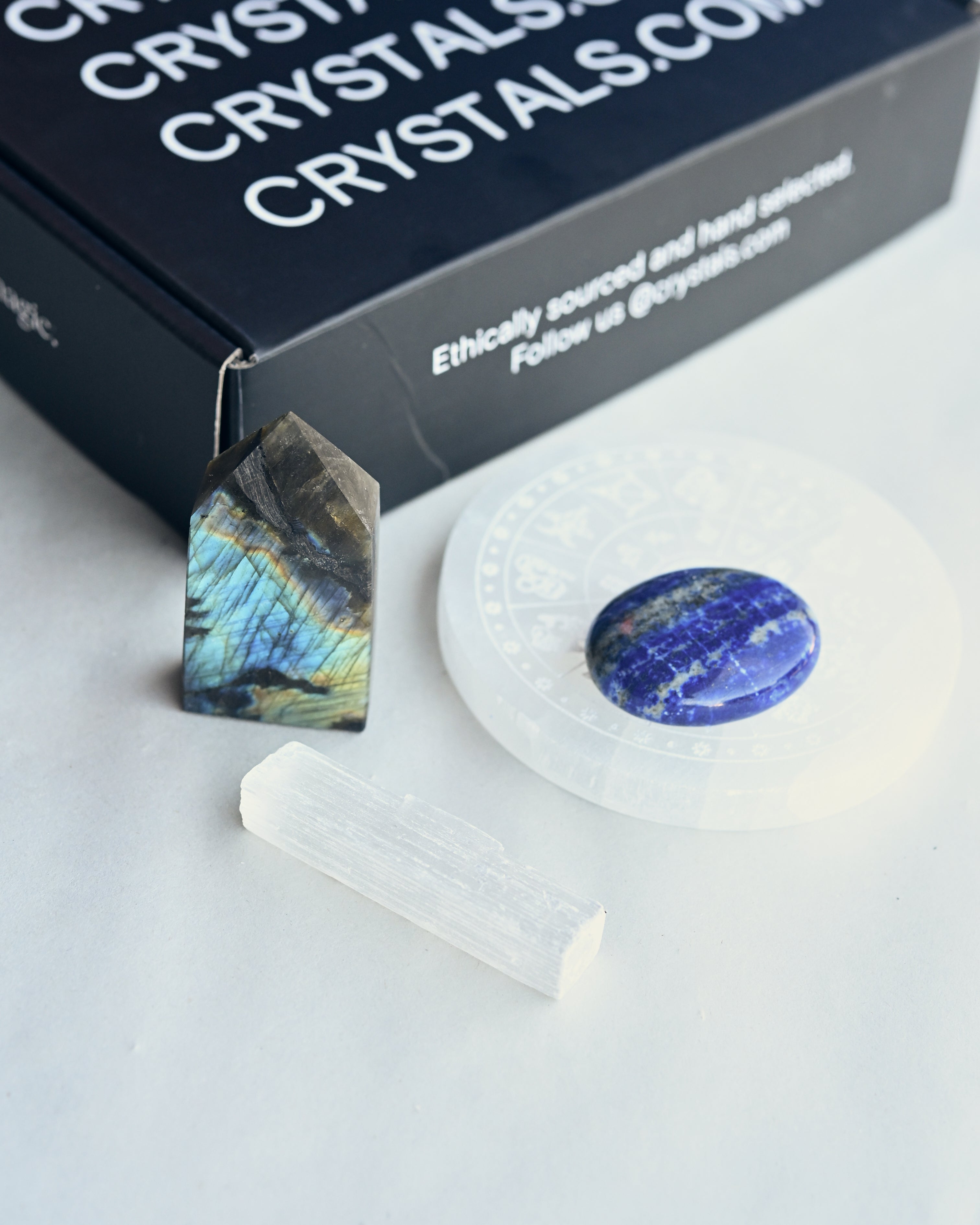 Crystal pendant, stone, and candle on a white surface with 'Crytal Crystals' branding.