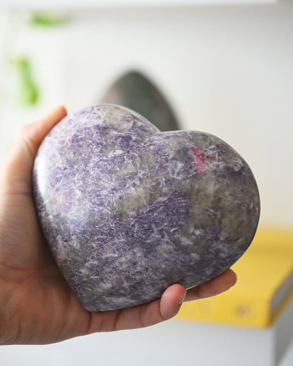 A hand holding a large heart-shaped Lepidolite crystal with light purple coloring.