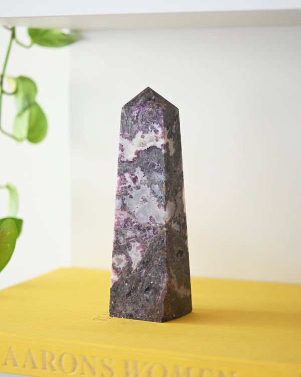 A polished Purple Agate Tower with deep violet and soft lavender hues, standing upright on a yellow surface with a plant in the background.