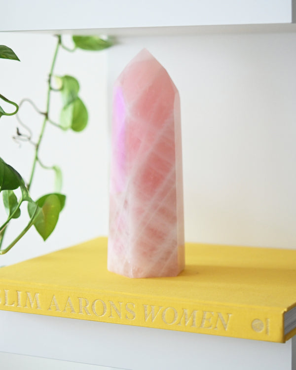 A large Rose Quartz Tower placed on a yellow surface with a book titled 'Women' visible in the background. The crystal is pink with visible natural patterns.