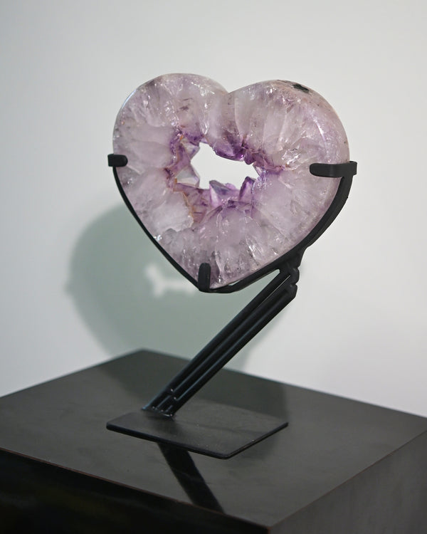 An amethyst crystal heart-shaped geode on a black custom stand, displaying a mix of purple and pink hues.