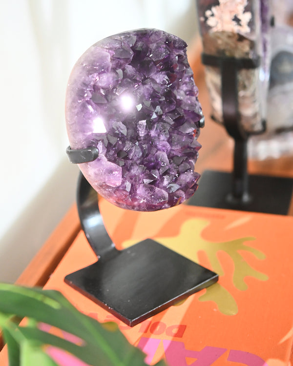 A large amethyst geode cluster on a custom black stand, displaying deep violet points and a radiant inner sparkle.