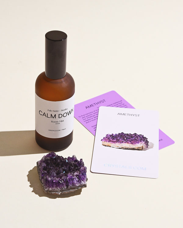A bottle of 'Calm Down Room Mist' with a dark label and a cluster of amethyst crystals on a light background, alongside a purple card with text.