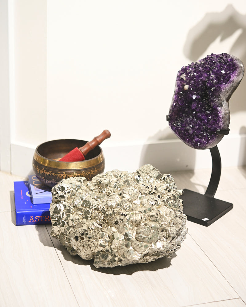 A large Pyrite cluster displayed on a stand, alongside a singing bowl, a box, and a crystal encrusted structure, all placed on a white surface.