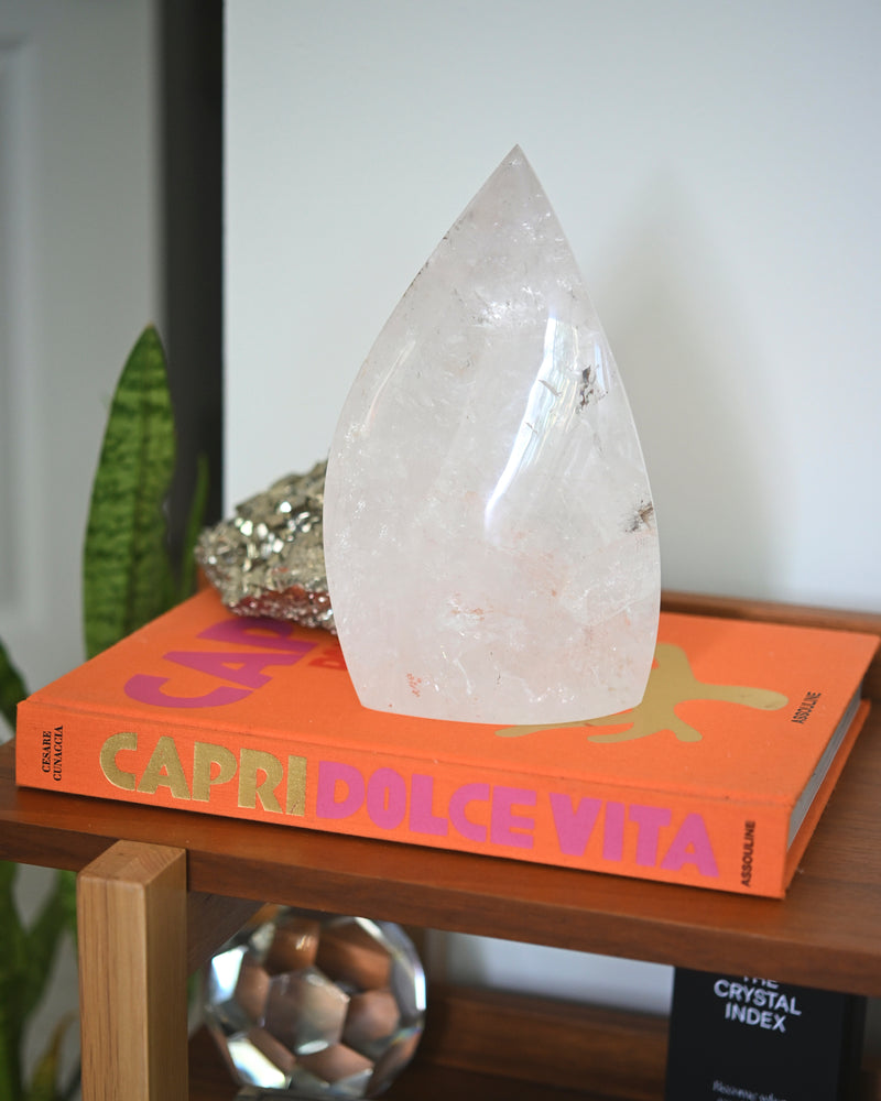 A large clear quartz crystal with golden rutile inclusions is displayed on top of an orange book titled 'CAPRIO DOLCE VITA'. The crystal is sculpted into a flame shape and occupies a prominent position in the image.